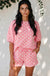 A woman modeling a pink two-piece lounge set with white polka dots, consisting of a short-sleeve top and matching shorts, set against a white wall with leafy vines and dappled sunlight.
