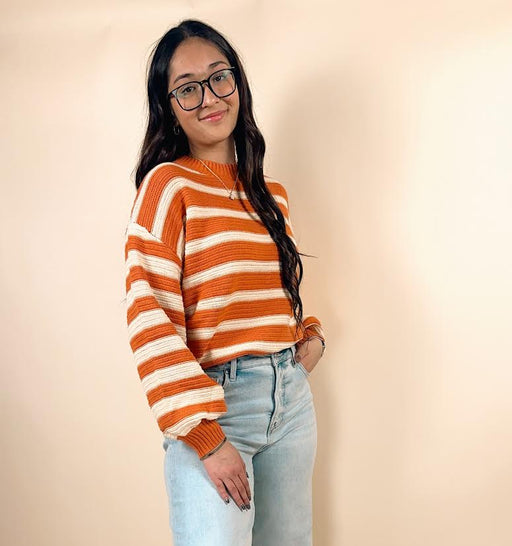 Person wearing an orange and white striped sweater with light blue jeans on a beige background