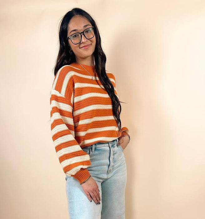 Person wearing an orange and white striped sweater with light blue jeans on a beige background