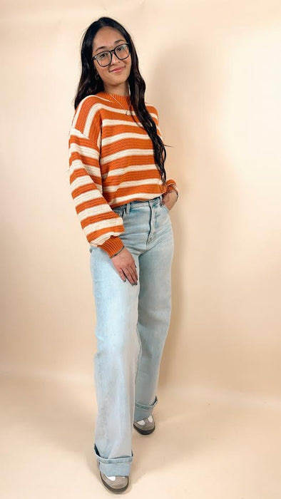 Woman wearing an orange and white striped sweater and light blue jeans against a beige background