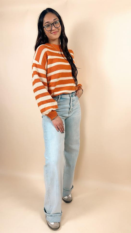 Woman wearing an orange and white striped sweater and light blue jeans against a beige background