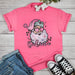 A pink t-shirt with a graphic of Santa Claus and the text 'SANTA BELIEVE' on the front, displayed flat with casual clothes and sneakers around it.