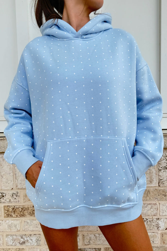 A person wearing an oversized light blue hoodie with a small white polka dot pattern, standing in front of a light brick wall.
